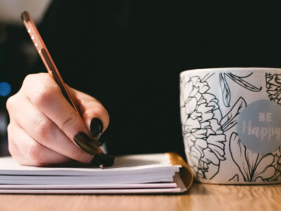 the hand of woman with black nail varnishhold an orange pen writing in an notebook. In the other hand she is holding a mug that says Be Happy - positive journal prompts