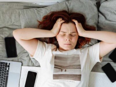 Picture of a frowning woman lying on a bed with her eyes closed and her hands in her hair. She is surrounded by a computer and several mobile phones