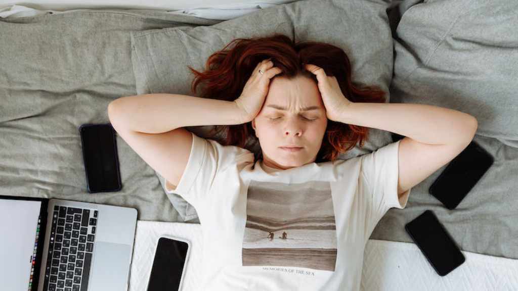 Picture of a frowning woman lying on a bed with her eyes closed and her hands in her hair. She is surrounded by a computer and several mobile phones