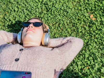 woman lying on her back on the grass. She's wearing sunglasses and listening to something from her phone through over ear headphones