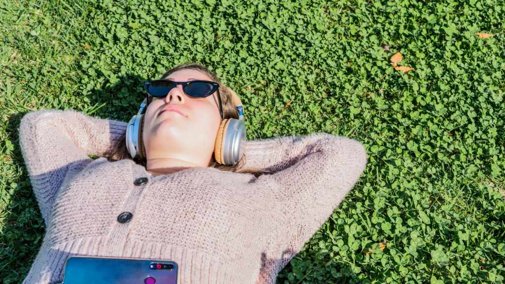 woman lying on her back on the grass. She's wearing sunglasses and listening to something from her phone through over ear headphones