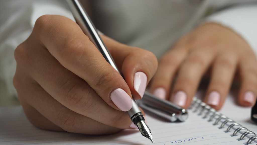 A woman with pink nail varnish writing in a notebook using a silver fountain pen