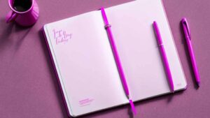A pink notebook and pen against a pink background with a pink coffee cup