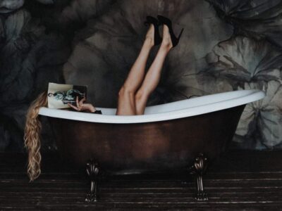 woman in a bath with her legs in the air and high heeled shoes on her feet, holding a magazine in front of her face