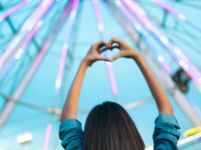 A woman stood looking at a ferris wheel with her hands lifted above her head making a heart shape