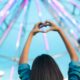 A woman stood looking at a ferris wheel with her hands lifted above her head making a heart shape