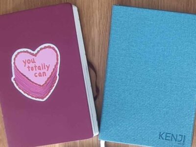 Two notebooks. One is purple with a heart shaped sticker that says you totally can. The other is blue and has Kenji embedded in the cover.