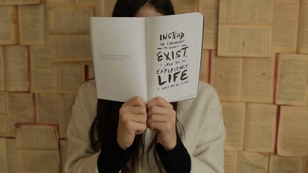 a woman holding up a book in front of her face. The page is open and it says Instead of choosing to merely exist, I urge you to experience life. I urge you to live. An example of a self help book for women