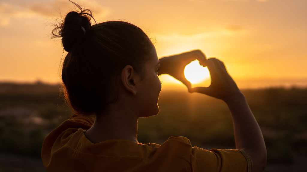 At sunset a women holding up her hands to make a heart shape around the sun