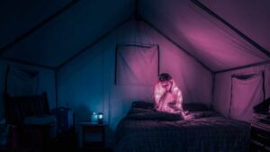 Girl sat on a bed in a tent. She has lights in her jumper which give the tent a pink glow which graduates to blue on the other side of the tent.